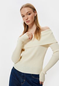 Leicht cremefarbener, schulterfreier, gerippter Strickoberteil mit langen Ärmeln und enganliegendem Design, kombiniert mit dunkelblauer Denim.