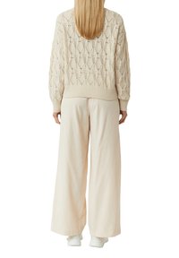 Gestrickter Pullover in Creme mit Rautenmuster, kombiniert mit weichen, weit geschnittenen hellbeigen Hosen. Weiße Schuhe runden das Outfit ab.