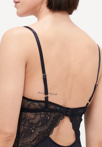 Fekete csipkebodysuit, állítható pántokkal, fodros szélekkel és kulcslyuk hátlappal, ami kapcsos záródású. Érdekessége egy bőrön látható tetoválás.