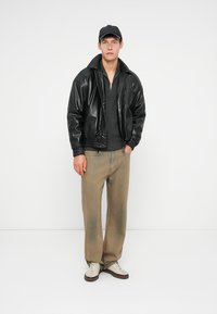 Černá kožená bomber bunda, šedý svetr na zip, béžové kalhoty a šedé tenisky. Model má na sobě černou čepici a stojí proti bílému pozadí.