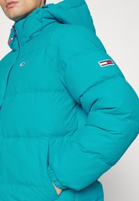 Teal puffjacka med huva, dragkedja framtill och elastiska ärmslut. Inkluderar en liten logotypspatch på vänster ärm.