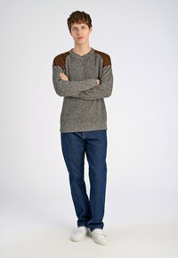 Suéter gris de punto con detalles en cuero marrón en los hombros, combinado con jeans azul oscuro y zapatillas blancas. El modelo está de pie con los brazos cruzados.