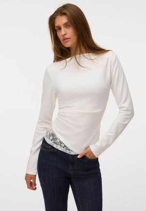 Jeune femme aux longs cheveux bruns portant un haut blanc à manches longues avec un détail en dentelle et un jean bleu foncé, posant avec une main dans la poche.