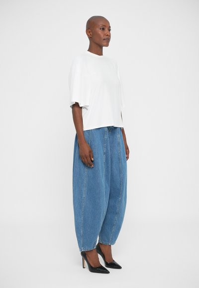 Vrouw die zijwaarts staat in een wit oversized T-shirt met korte mouwen, brede blauwe jeans en zwarte hoge hakken tegen een witte achtergrond.