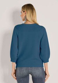 Frau mit langen blonden Haaren trägt einen blauen Strickpullover mit 3/4-Ärmeln und grauen Jeans, von hinten vor einem schlichten Hintergrund gezeigt.