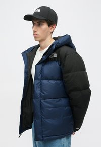 Mornarska plava i crna jakna s kapuljačom, s teksturiranim završetkom i vodoravnim prošivanim uzorkom. Logo na prsima, zatvarač.