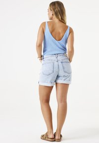 Top sin mangas de color azul claro con espalda en V, combinado con pantalones cortos de mezclilla clara de cintura alta, con dobladillos enrollados, y zapatos slip-on marrones. Fondo neutro.