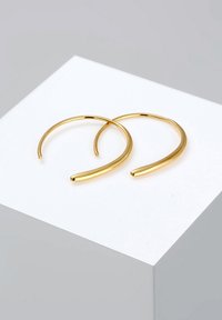 Boucles d'oreilles en métal doré en forme de hoops, avec un design lisse et courbé et des extrémités effilées, reposant sur une surface d'exposition blanche.