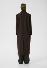 Manteau long marron à coupe droite, grand col et poches latérales fendues. Porté sur un pantalon noir à jambes larges. Texture en laine.