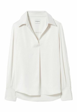 Femme debout face à l’objectif, portant un chemisier blanc à col, un pantalon blanc large, une ceinture à imprimé léopard et des sandales compensées noires avec des détails tissés.