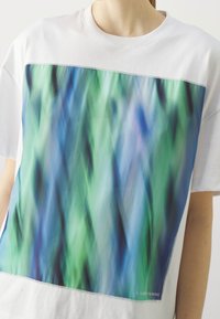 Camiseta blanca con un gran gráfico frontal en tonos de verde y azul, que presenta un diseño abstracto borroso. Mangas cortas, cuello redondo.