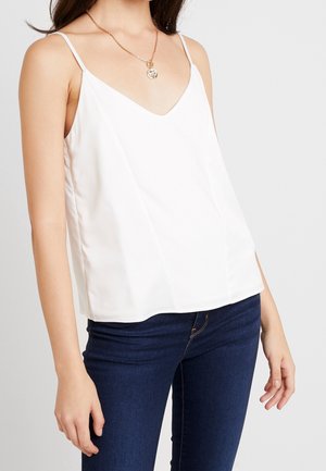 Kobieta ubrana w biały, bezrękawnik z dekoltem w serek oraz ciemnoniebieskie dżinsy, z złotym naszyjnikiem z zawieszką, stoi na tle jasnego, jednolitego tła.