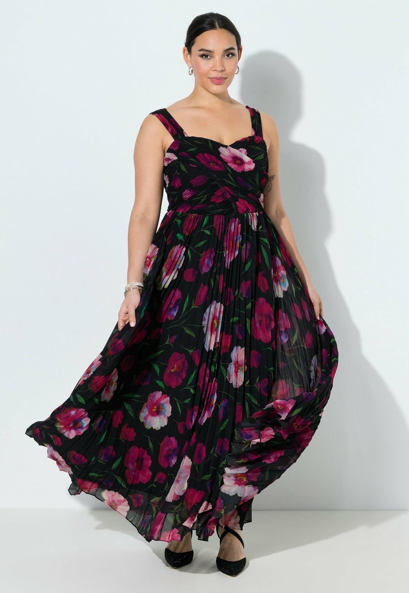 Mujer con vestido maxi negro sin mangas con estampado floral rojo y rosa, sosteniendo la falda, llevando tacones negros y pulsera plateada.