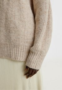 Maglione lavorato a maglia beige chiaro con texture a coste, caratterizzato da una vestibilità ampia e maniche lunghe. Indossato sopra una gonna plissettata color crema.