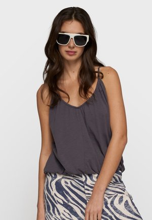 Donna con lunghi capelli scuri che indossa occhiali da sole neri con montatura bianca, top grigio scuro senza maniche e pantaloni beige con strisce astratte zebrate blu.