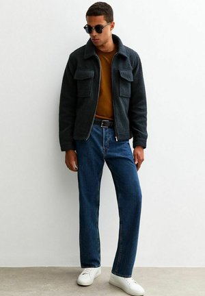 Schwarze Fleecejacke mit zwei frontalen Taschen, braunem Hemd darunter, blauen Jeans und weißen Sneakers, getragen von einer Person mit Sonnenbrille.