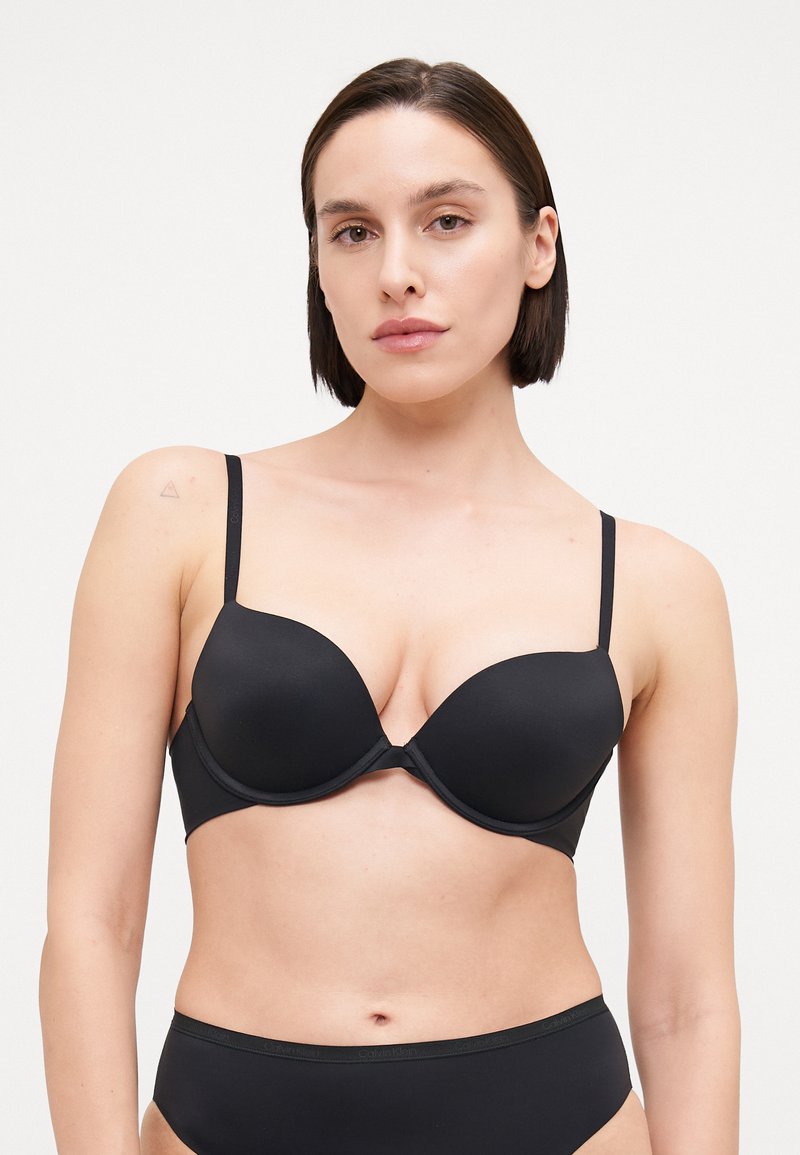 Svart push-up BH med glatte, formede kopper, justerbare stroppar og eit sømlaus design. Materialet verkar mjukt med ei matt finish.