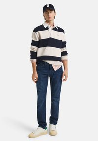 Maglione a righe navy e crema sopra una camicia rosa con colletto, abbinato a jeans blu scuro e sneakers bianche con dettagli verdi.