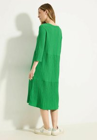 Grün, strukturiertes Midi-Kleid mit Dreiviertelärmeln und gestufter Gestaltung. Kombiniert mit weißen Sneakers mit grünen Akzenten.