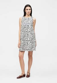 Γυναίκα με μακριά σκούρα μαλλιά, φοράει αμάνικο ασπρόμαυρο φόρεμα με λουλούδια και σκουροκόκκινα παπούτσια Mary Jane, στέκεται σε απλό λευκό φόντο.