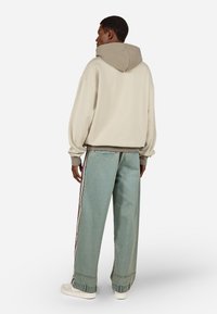 Ανωμί γυαλιστερό hoodie σε ανοιχτό μπεζ χρώμα με καπιτονέ καπέλο σε αντίθεση καφέ χρώμα, συνδυασμένο με ελαφριά τζιν παντελόνια σε ψηλόμεσο φαρδύ κόψιμο, που διαθέτουν κόκκινες λωρίδες στο πλάι.