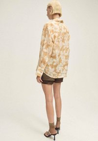 Camicia a maniche lunghe in beige chiaro con motivo floreale bianco, indossata con pantaloni corti marroni. La camicia presenta una vestibilità rilassata e polsini con bottoni.