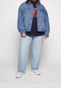 Osoba w jasnoniebieskiej dżinsowej kurtce, granatowej bluzie z czerwonym napisem, jasnoniebieskich dżinsach i białych trampkach, stojąca na jednolitym tle.