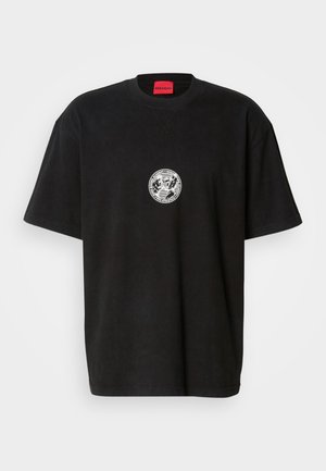 Tricou negru oversized cu mâneci scurte, o grafică mică rotundă albă pe piept și un eticheta roșie în guler care scrie "HUGO."