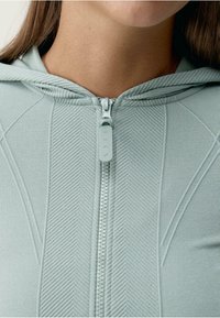 Svijetlozeleni zip-up dukserica koja sadrži teksturirani materijal, reljefni riblji uzorak i odgovarajući zip, s opuštenim krojem.