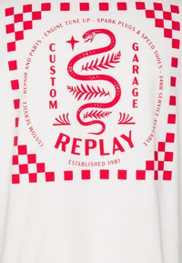 Bílá tričko s červeným grafickým designem, na kterém je had a kostkovaný vzor. Text obsahuje "CUSTOM GARAGE" a "REPLAY," založení 1981.