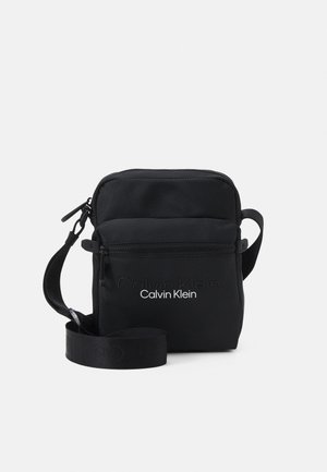 Mała czarna torba na ramię Calvin Klein z przednią kieszenią na suwak i regulowanym paskiem na jasnym tle.