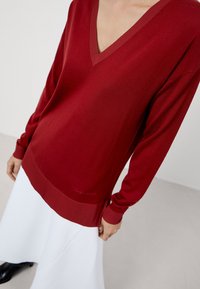 Rode trui met lange mouwen, V-hals, geribbelde halslijn en zoomdetail. Zachte textuur, iets losse pasvorm, gecombineerd met een witte rok.