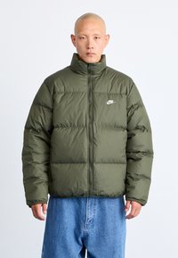 Olive green puffer jacket with a high collar, zip closure, and white Nike logo on the chest. Features a quilted design and roomy fit.