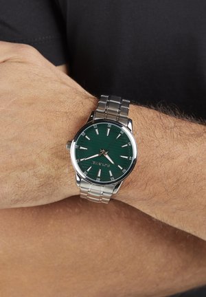 Zilverkleurig horloge van roestvrij staal met een groen gestructureerde wijzerplaat, zilveren uurmarkeringen en een datumindicator op 3 uur, gedragen om de pols.