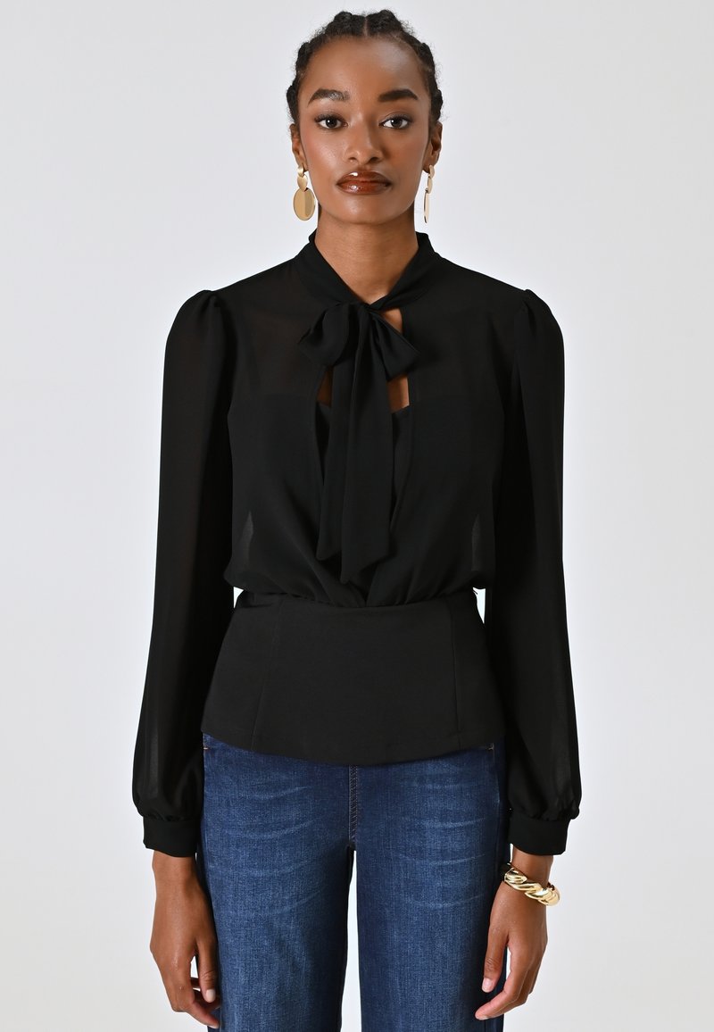 Schwarze Bluse mit einer Schleife am Hals, Puffärmeln und taillierter Taille. Getragen mit blauen Jeans. Glattes Material, minimales Muster.