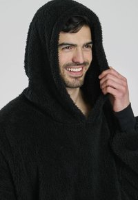 Felpa nera in pile con grande cappuccio, texture morbida e maniche lunghe. Modello sorridente mentre sistema il cappuccio con una mano.
