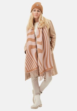 Hellbrauner Wollschal mit wellenförmigen pinken Mustern und Fransen, kombiniert mit einem beigen Mantel, einer cremefarbigen Strickjacke, hellen Hosen und weißen Stiefeln.