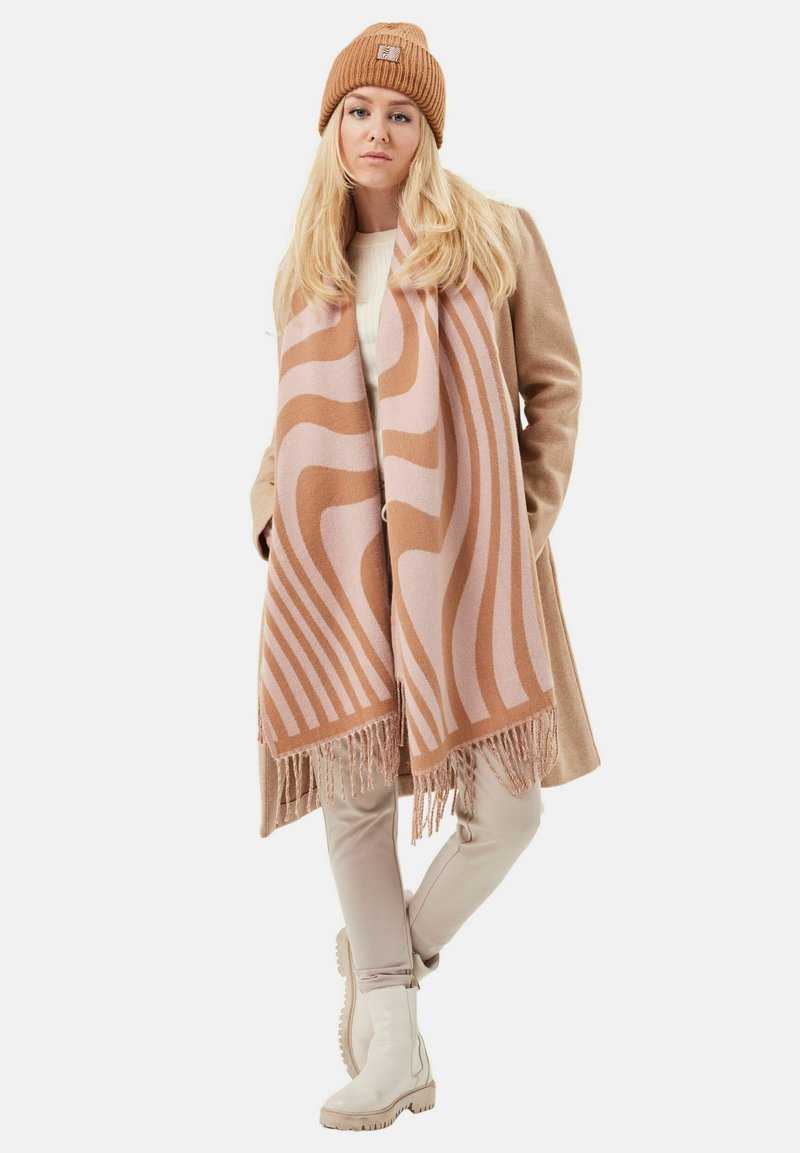 Tan wool scarf with wavy pink patterns, fringed edges, paired with a beige coat, cream sweater, light pants, and white boots.