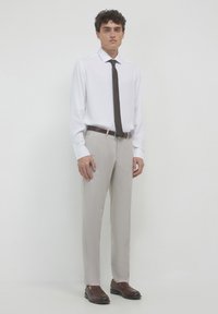 Homme portant une chemise habillée blanche à manches longues, une cravate marron foncé, un pantalon beige, une ceinture marron et des chaussures habillées en cuir marron, se tenant devant un fond blanc.