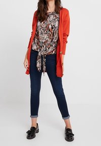 Oranje lange cardigan over een gestreepte blouse met een tailleband, gedragen met donkere skinny jeans en zwarte loafers met strikaccenten.