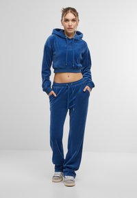 Felpa corta in velluto blu con lacci, abbinata a pantaloni jogger coordinati con vita elastica e tasche laterali. Sneakers grigie.