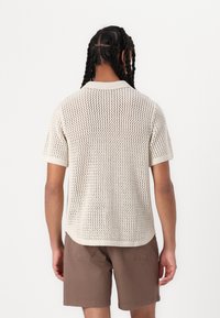 Pull en maille à manches courtes de couleur beige clair, doté d’un design en maille lâche, d’un col côtelé et d’un ourlet légèrement arrondi, porté avec un short marron.