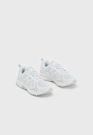 Para białych sportowych butów do biegania z siateczkowymi cholewkami i teksturowanymi gumowymi podeszwami, ułożonych obok siebie na jasnym tle.