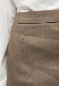 Gros plan sur la taille d'une jupe marron ajustée avec une couture latérale, portée avec une blouse blanche rentrée dedans.