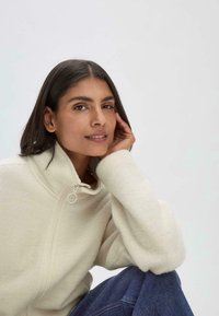 Cremfarbene Fleece-Jacke mit Reißverschluss und hohem Kragen sowie weicher Textur, ausgestattet mit einem runden Reißverschluss-Zug. Das Modell sitzt und trägt Jeans.