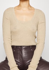 Haut beige côtelé à manches longues avec un col carré, présentant un effet subtil de scintillement. Associé à un pantalon en cuir marron foncé.