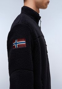 Schwarze Fleecejacke mit Reißverschlusskragen und sichtbaren Nähten, versehen mit einem roten Patch mit einer blau-weißen norwegischen Flagge am linken Ärmel.