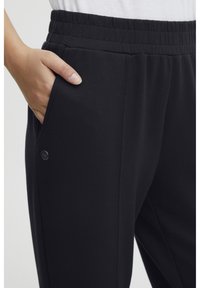 Sorte joggebukser med elastisk liv, sidelommer og en knappdetalj på lommen. Laget av et mykt, teksturert materiale.
