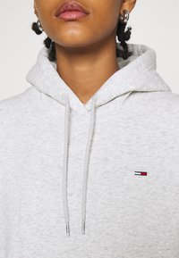 Grauer Kapuzenpullover mit Kordelzügen, aus weichem Stoff gefertigt. Verfügt über ein kleines gesticktes Logo mit roten, weißen und blauen Streifen auf der Brust.