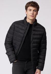 Giacca a vento nera con colletto alto, caratterizzata da cuciture orizzontali e chiusura con zip. Il materiale sembra liscio e leggero.